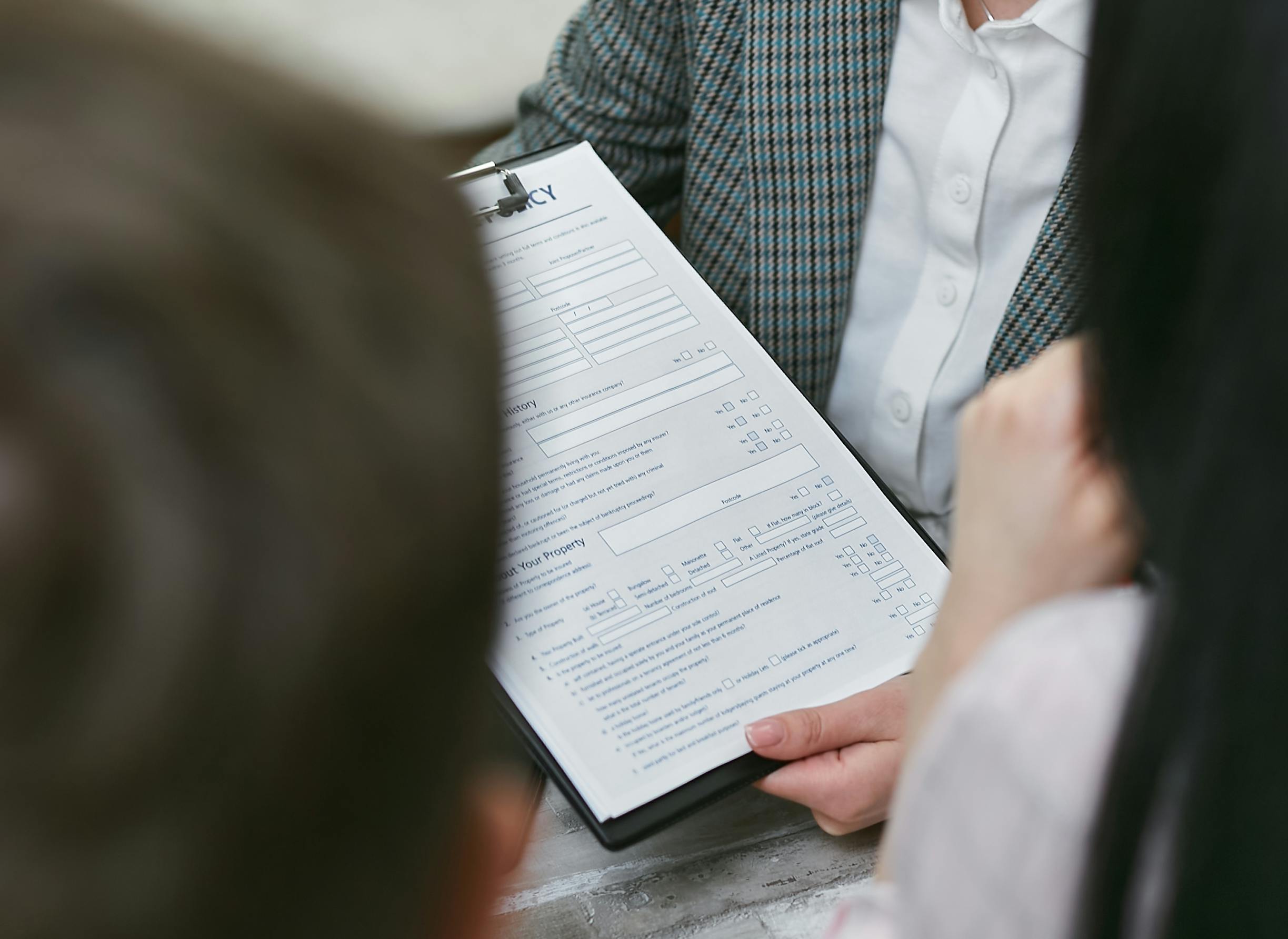 A person in business attire holds a clipboard with a detailed form, reviewing it with two individuals seated across the table.