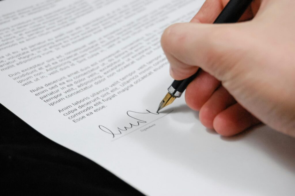 A hand signing a printed document with a black fountain pen, featuring blocks of text and a visible signature line.