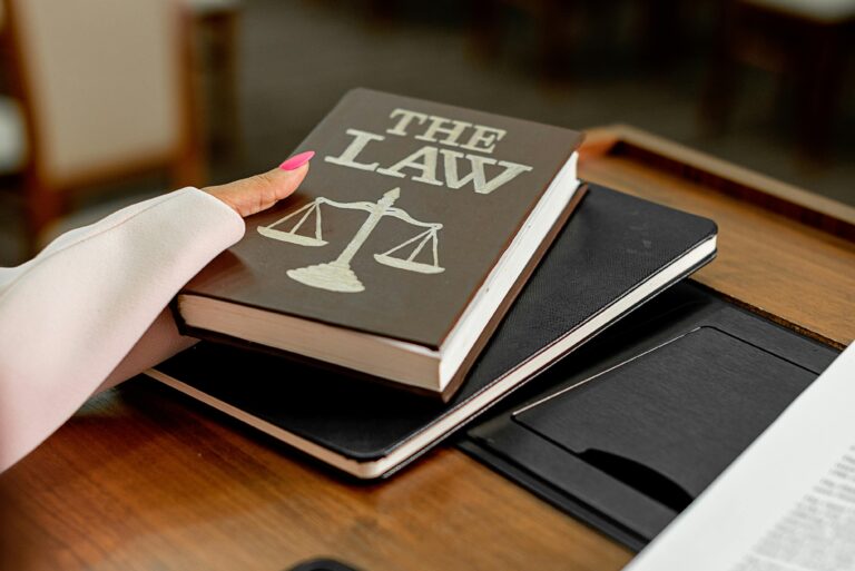 A person with pink nails holds a law book with a justice scale symbol, resting on a notebook on a wooden desk.