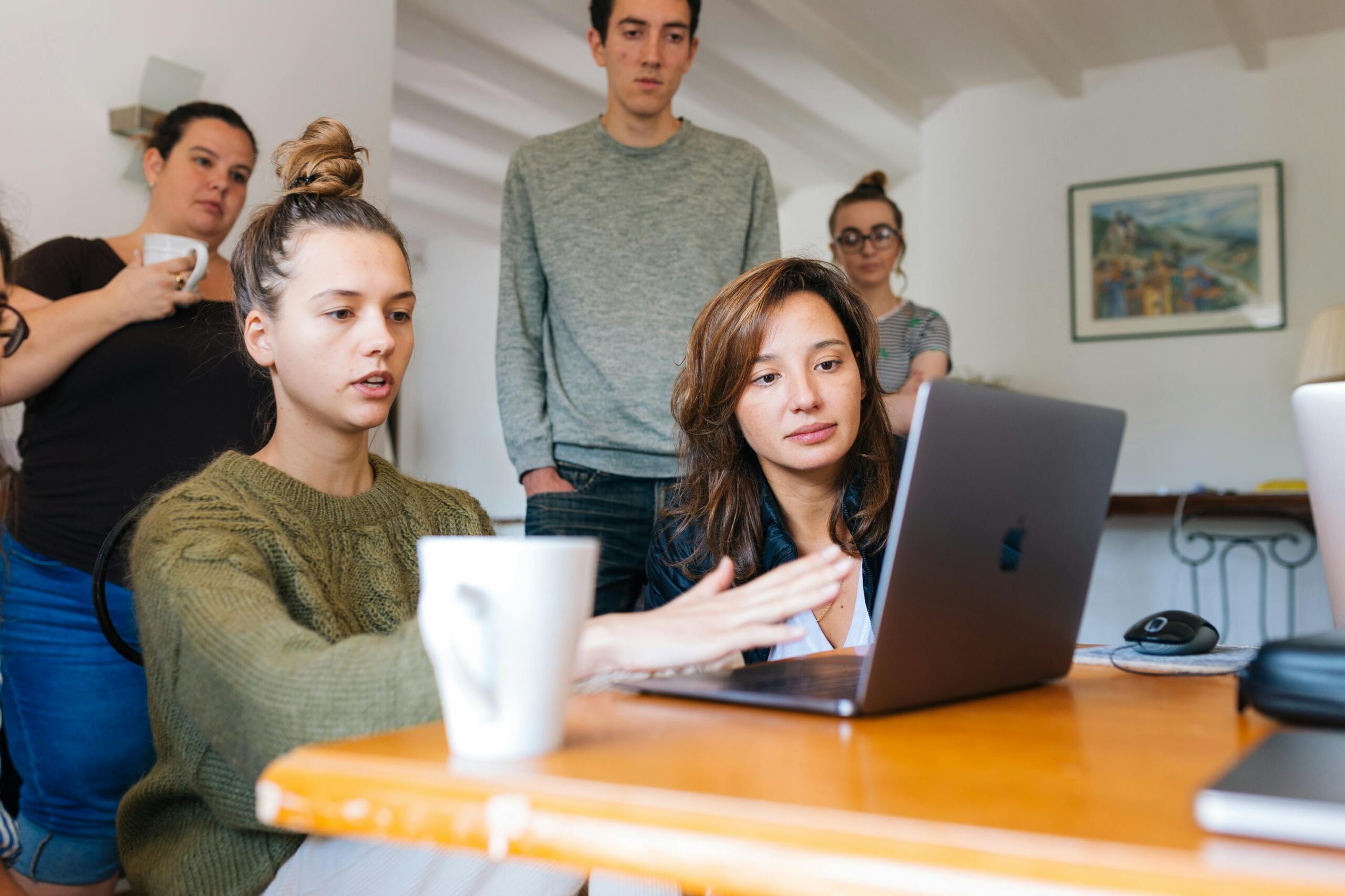 A group of young professionals gathered around a laptop in a casual office setting, engaged in a collaborative discussion.