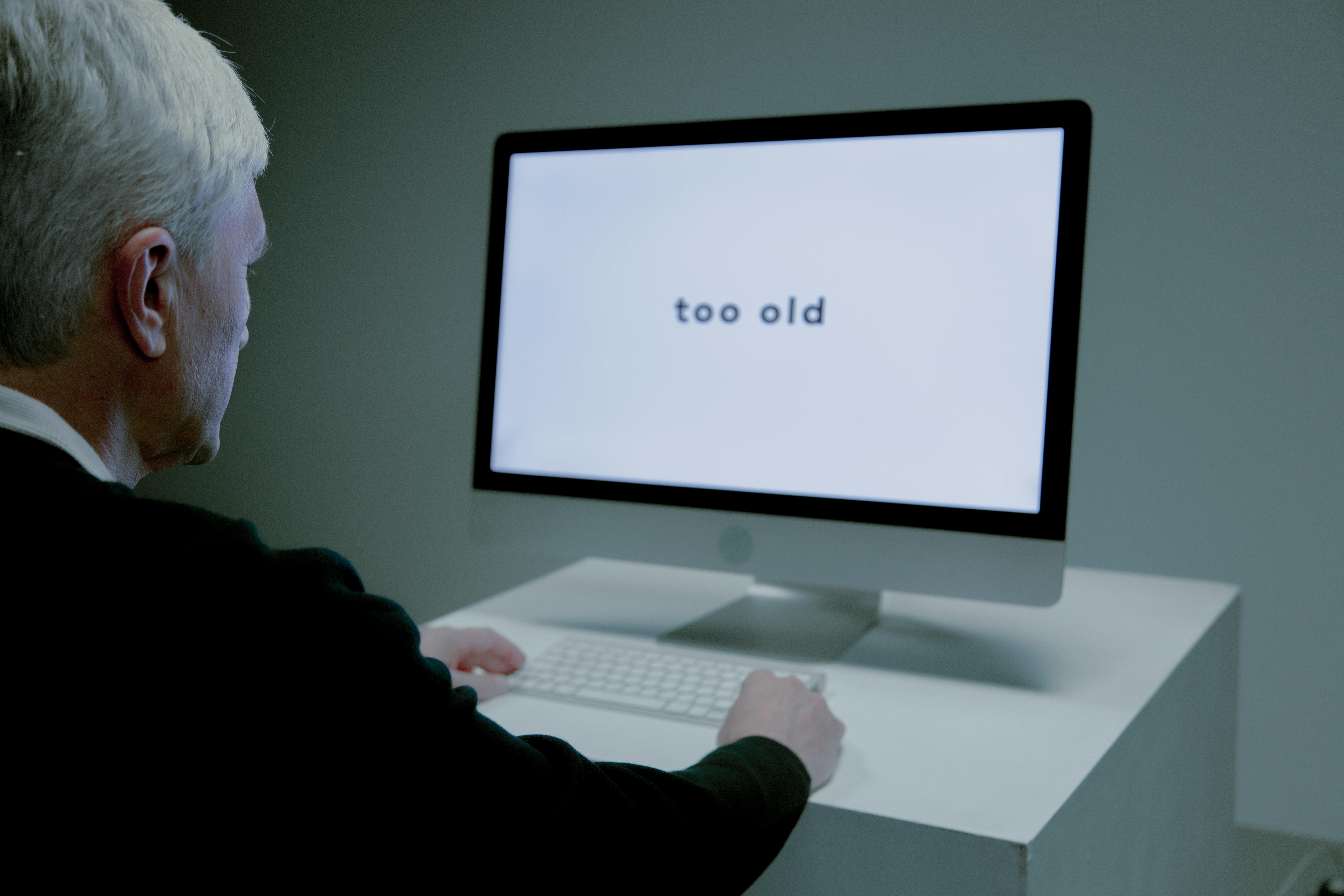 An older man sits at a computer displaying the words "too old," highlighting a potential case of age discrimination in the workplace.