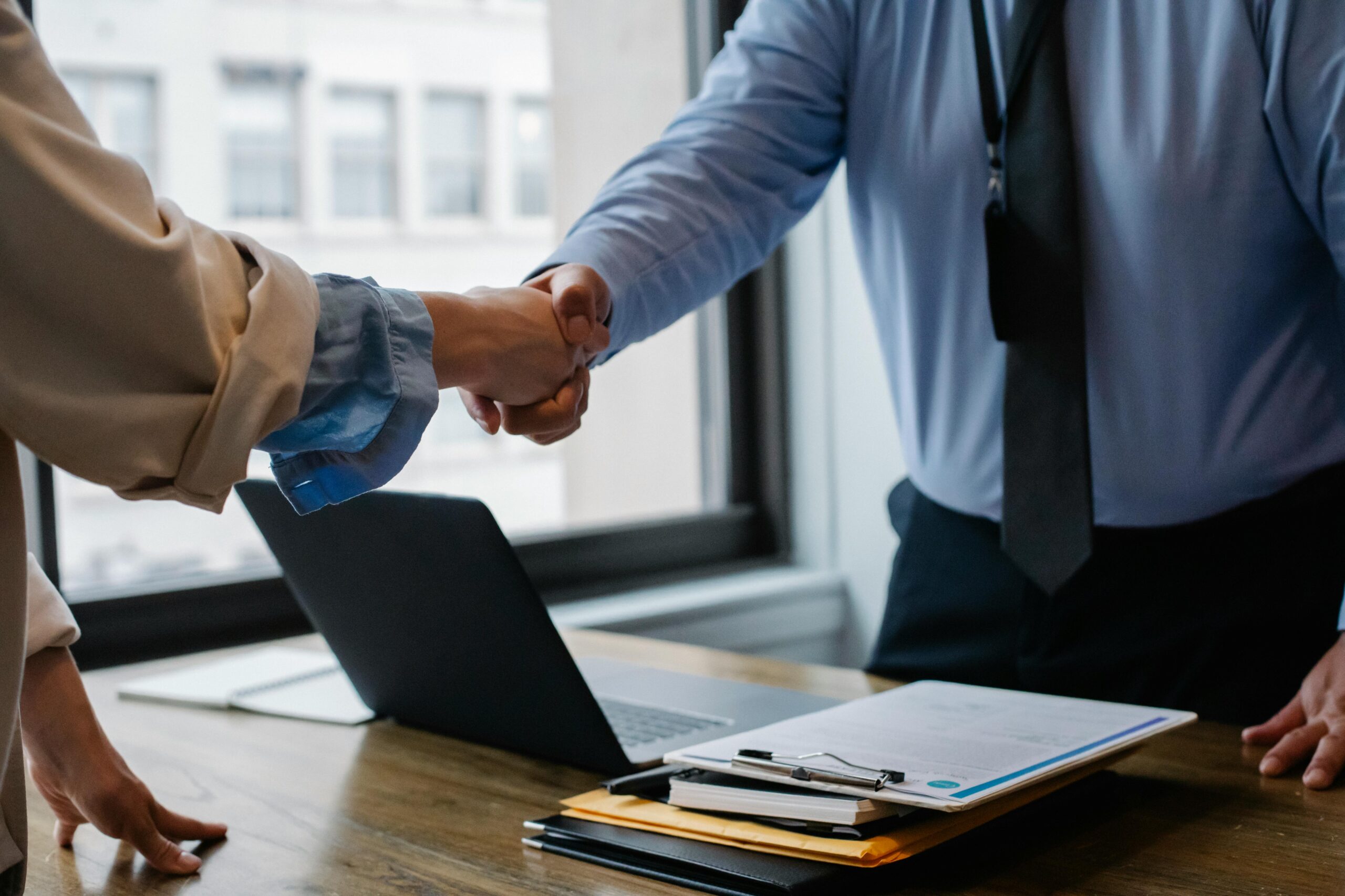 Two professionals shake hands over a desk with documents, a laptop, and office supplies, symbolizing agreement or partnership.
