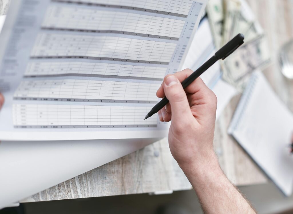 a person holding a pen and reviewing a printed spreadsheet or timesheet. In the background, there are dollar bills, a notebook, and papers on a wooden surface—suggesting themes like payroll, compliance, or financial documentation.